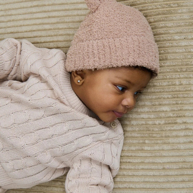A baby wearing a cream cable-knit sweater and a dusty pink knit beanie hat, lying on a ribbed beige blanket while holding an adult's hand.