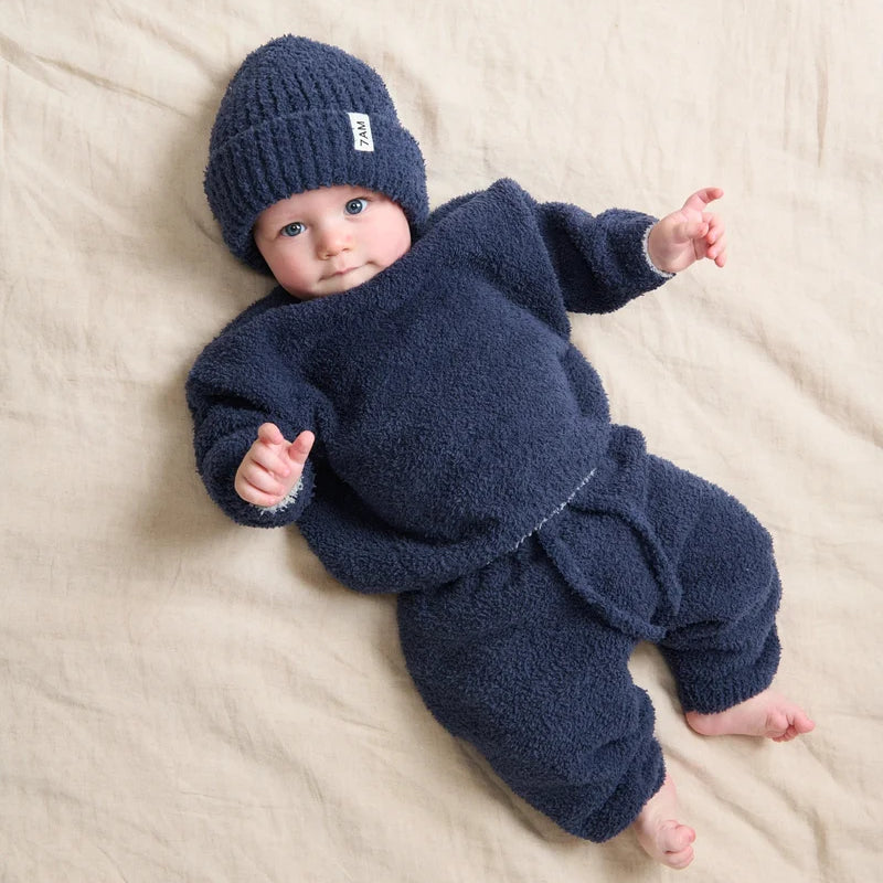 A baby wearing a dark navy blue ribbed knit beanie and a matching fuzzy long-sleeved outfit while lying on a soft, light-colored surface.
