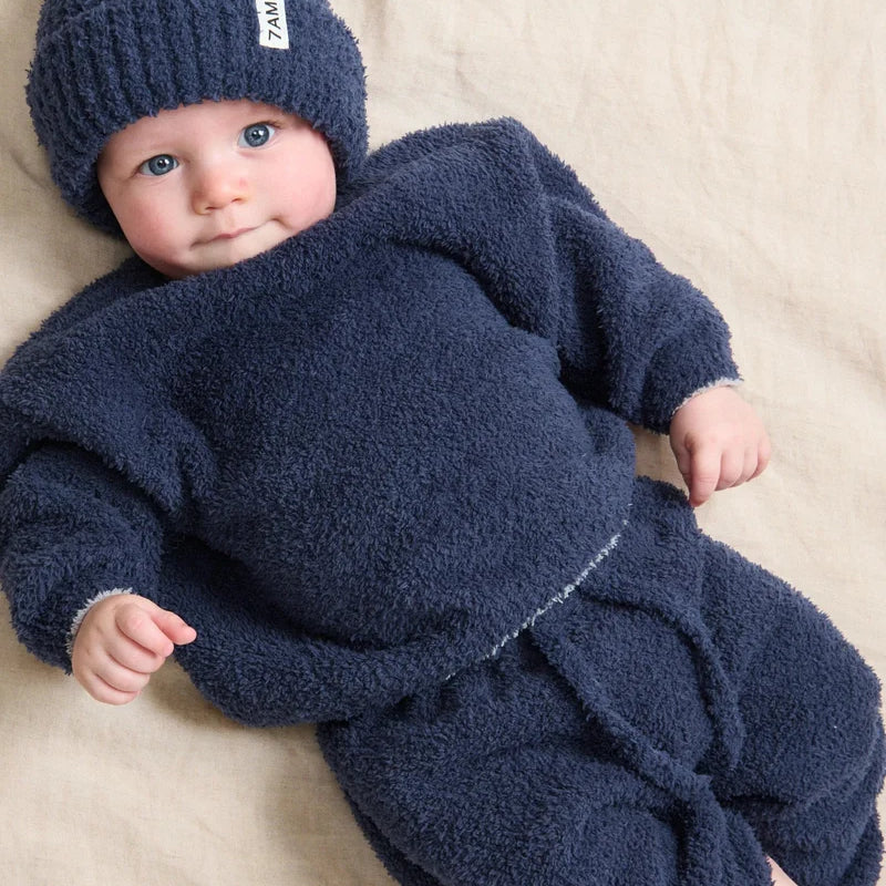 A baby lying down and wearing a dark navy blue ribbed knit beanie with a small white brand tag and a matching fuzzy long-sleeved outfit.