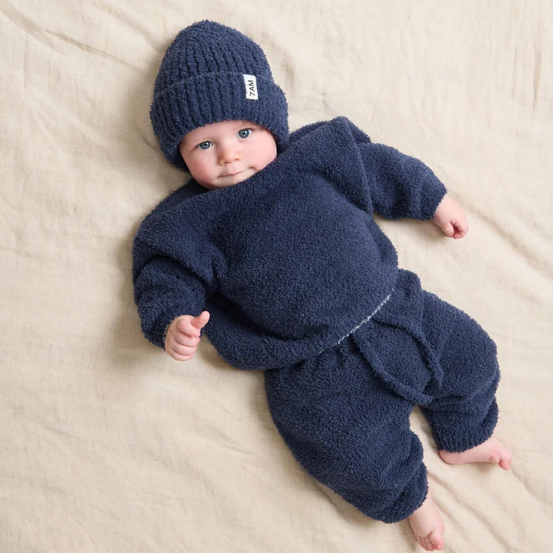 A baby lying on a beige fabric surface wearing a matching navy blue fuzzy knit outfit with a beanie hat. A small white tag on the hat reads 'BEBE'.