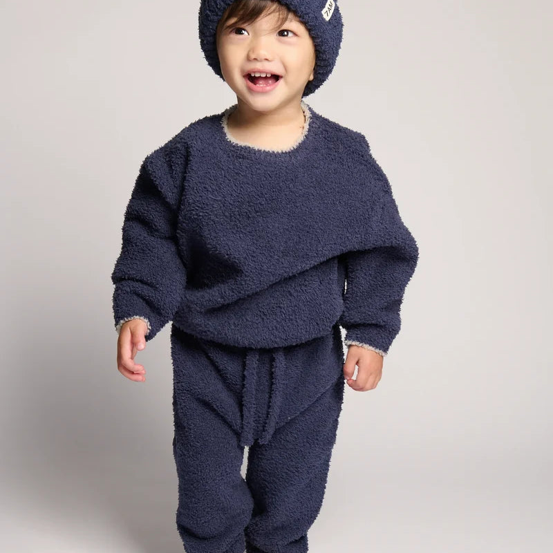 A smiling toddler wearing a matching navy blue fuzzy knit outfit, including a beanie, long-sleeve top, and pants, standing against a light gray studio background.