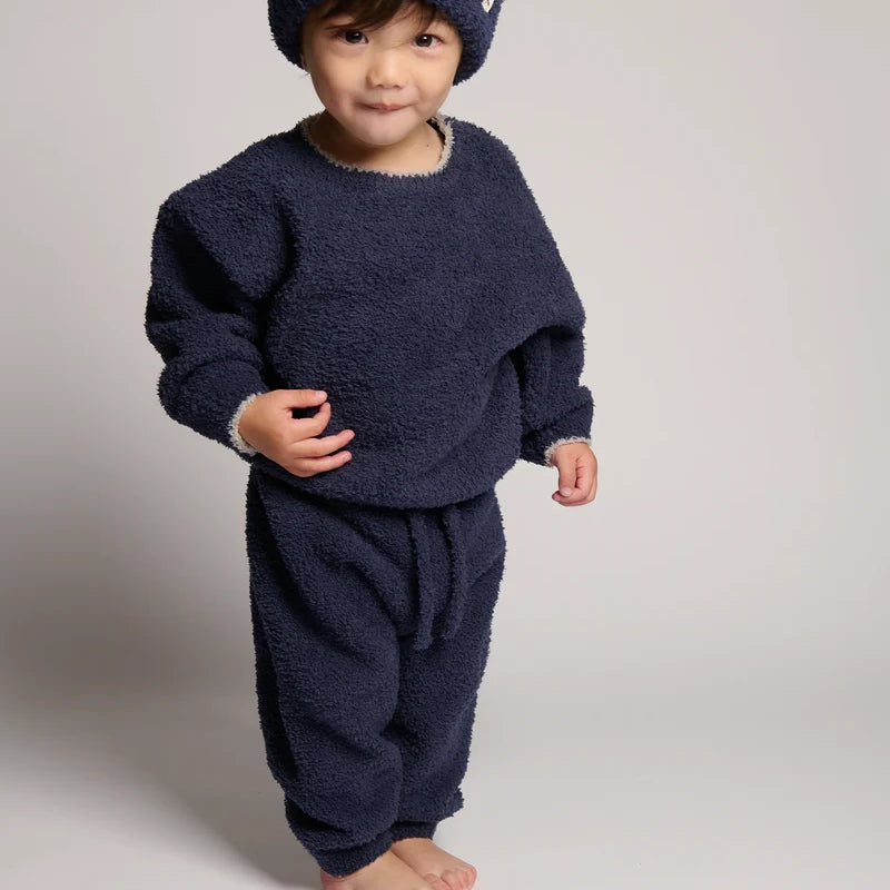 A toddler wearing a cozy navy blue fuzzy knit outfit, including a matching beanie, sweatshirt, and pants, photographed against a neutral light gray studio background.
