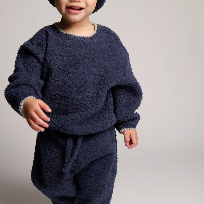A smiling toddler wearing a matching navy blue fuzzy fleece outfit with a knit beanie, photographed against a plain light gray studio background.