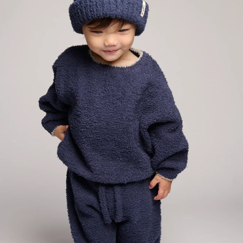 A toddler wearing a cozy navy blue fuzzy knit outfit, including a matching beanie, sweatshirt, and pants, photographed against a neutral light gray studio background.