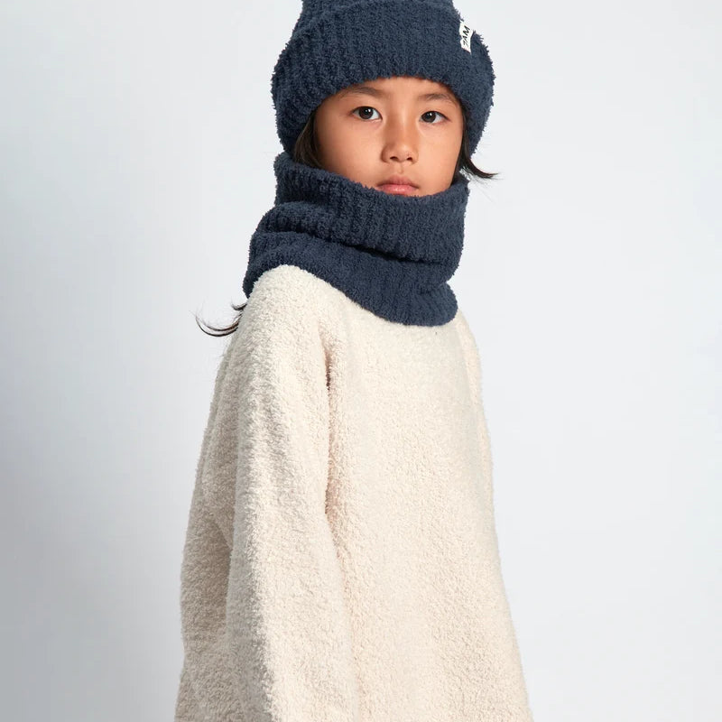 A child wearing a navy blue knit beanie with a small white logo and a matching navy blue knit neck warmer, paired with a cream sherpa sweater, against a light gray studio background.