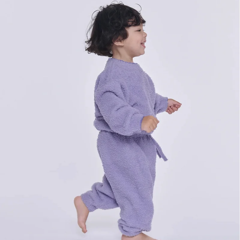 A toddler walking sideways against a light gray background, wearing a fuzzy lavender long-sleeve sweatshirt and matching lounge pants.