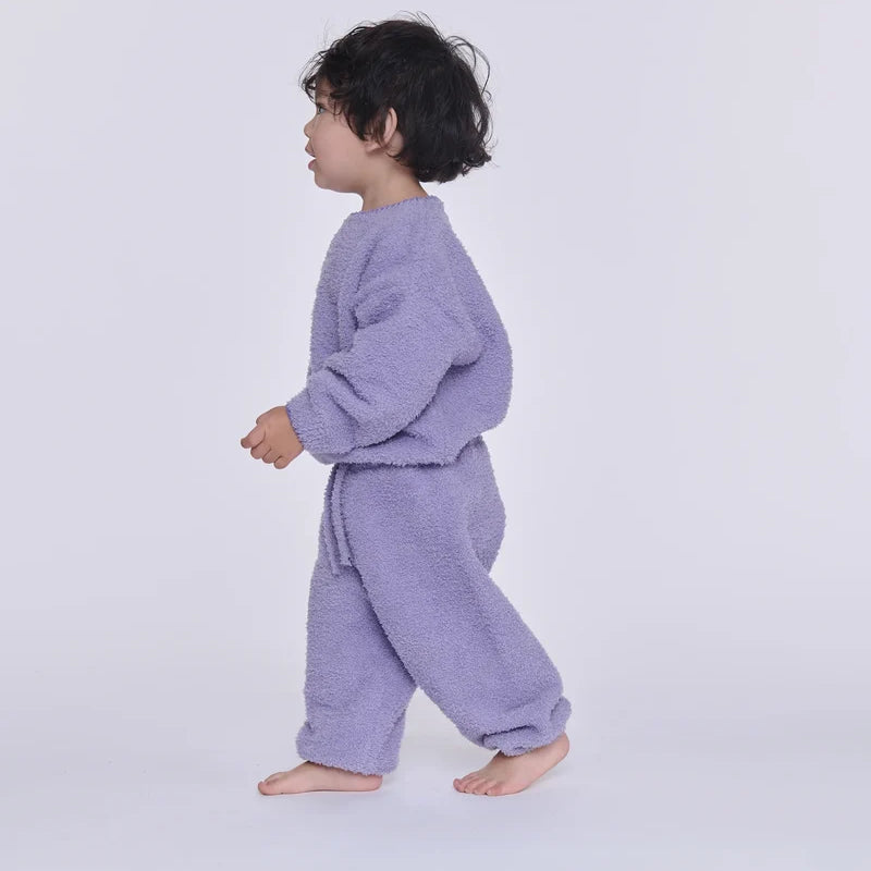 A toddler walking in profile wearing a fuzzy lavender two-piece lounge set against a plain light gray studio background.