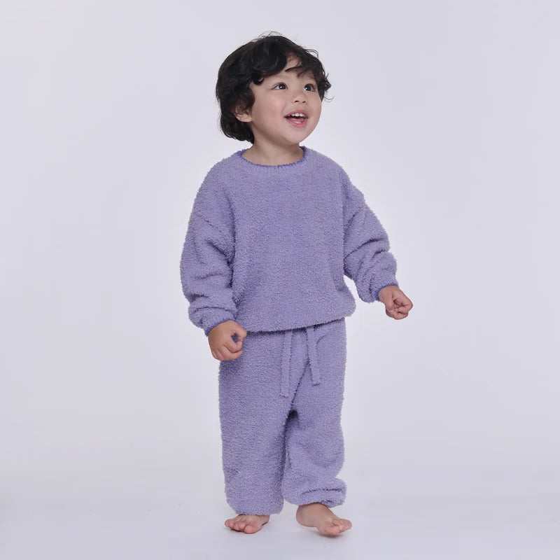 A toddler wearing a fuzzy lavender two-piece loungewear set, consisting of a long-sleeve sweatshirt and matching pants, standing against a plain light gray background.