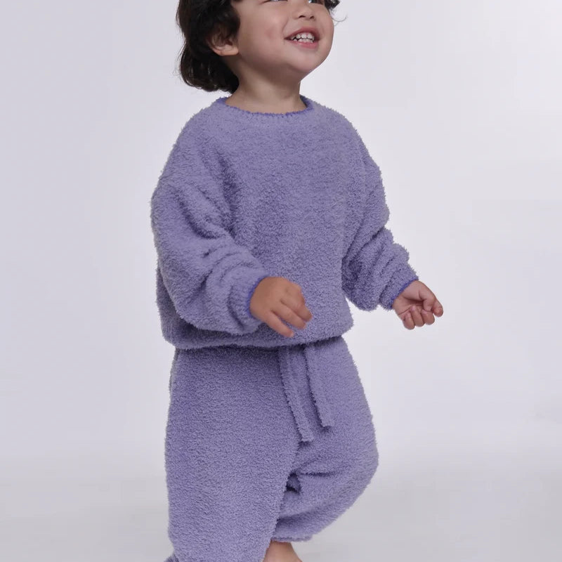 A toddler wearing a fuzzy lavender two-piece loungewear set, consisting of a long-sleeve sweatshirt and matching pants, standing against a plain light gray background.