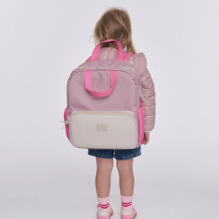 A young child viewed from behind wearing a dusty pink puffer jacket, denim shorts, and pink shoes, carrying a large structured diaper bag backpack in dusty pink and cream. The backpack features bright pink handles, side pockets, and a cream front pocket with a small metallic brand plaque.