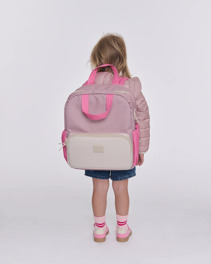A young child viewed from behind wearing a dusty pink puffer jacket, denim shorts, and pink shoes, carrying a large structured diaper bag backpack in dusty pink and cream. The backpack features bright pink handles, side pockets, and a cream front pocket with a small metallic brand plaque.