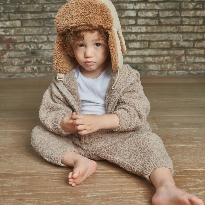 Child wearing a beige fleece outfit with a matching hat sitting on a wooden floor against a brick wall.