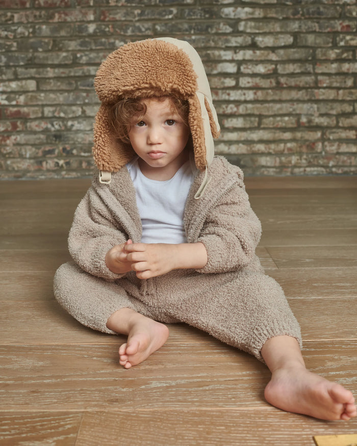 Child wearing a beige fleece outfit with a matching hat sitting on a wooden floor against a brick wall.