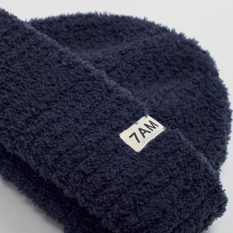 A close-up of a dark navy blue fuzzy fleece beanie with a small white rectangular label reading '7AM' on the cuff, set against a light gray background.