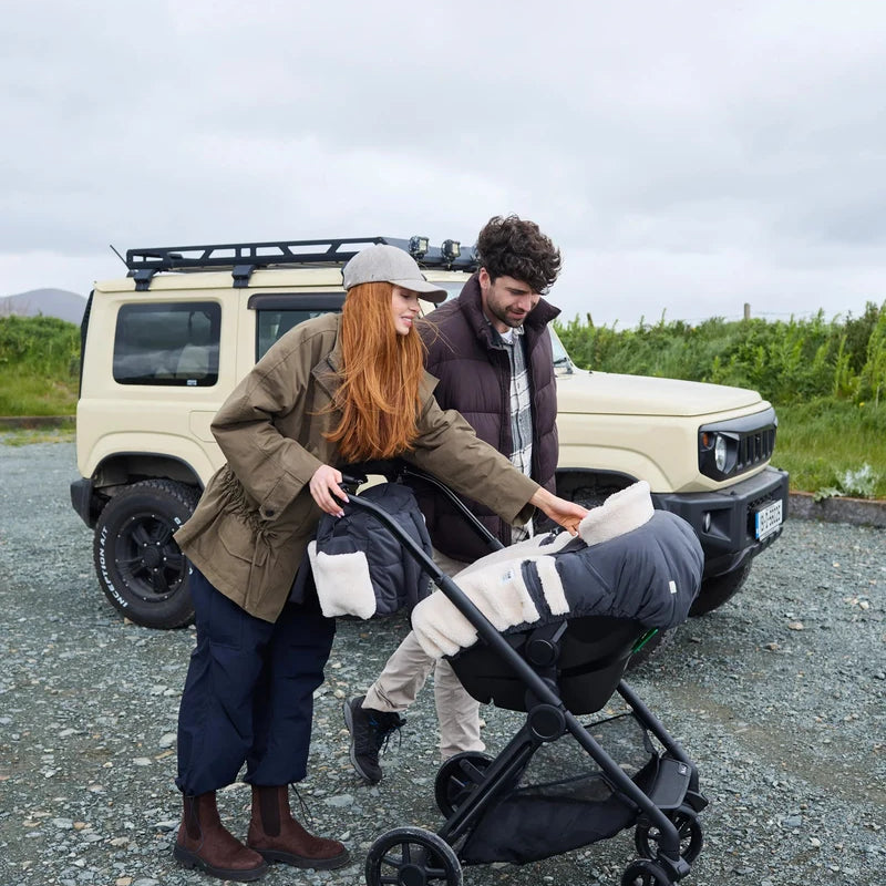 A woman pushes a stroller with a baby inside while walking outdoors next to a man in front of a beige off-road vehicle under an overcast sky, with the baby carriage outfitted with a Benji Applique Car Seat Cocoon by 7AM Enfant.