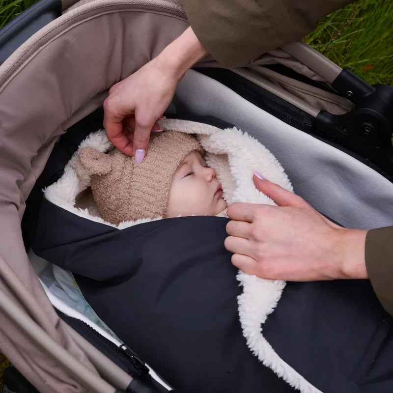 An adult adjusting a stroller bag with white fleece lining while a baby wearing a bear-eared knit beanie rests inside the stroller.