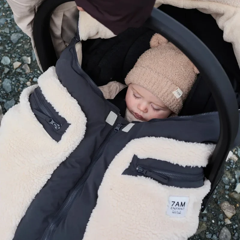 A baby sleeping in a black stroller wearing a beige knit beanie, covered by a cream and black fleece-lined stroller bag with zippered pockets. A small white label on the bag reads '7AM ENGLAND'.