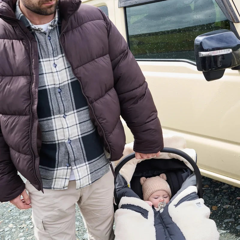 A man standing next to an SUV outdoors, holding a baby in a stroller who is wearing a fuzzy bear-eared beanie and a warm winter bunting bag.