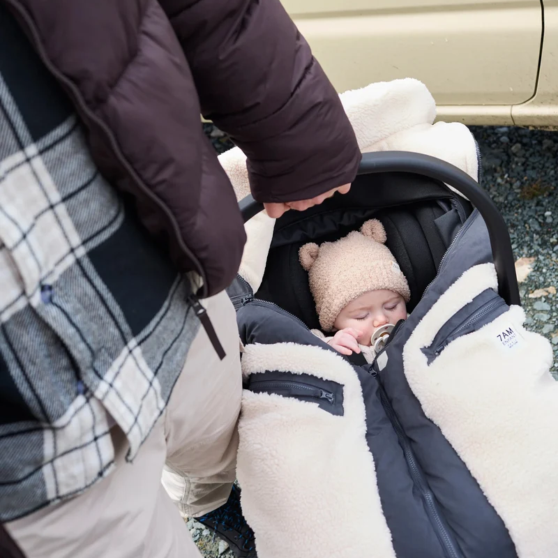 A baby wearing a beanie and pacifier is snuggled inside an infant car seat with the Benji Applique Car Seat Cocoon by 7AM Enfant featuring a dark navy or black outer shell and ivory sherpa trim, while a person stands nearby holding the car seat handle.