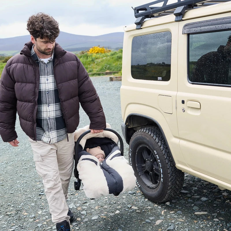 A man walking beside a cream-colored off-road vehicle, carrying a baby in a padded car seat. The man wears a dark brown puffer jacket over a plaid shirt and beige pants, set against a gravel ground and distant hills.