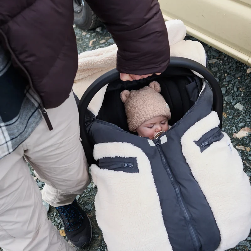 A baby sleeping in a stroller wearing a beige knit beanie, covered by a cream and black fleece-lined stroller bag with zippered pockets.