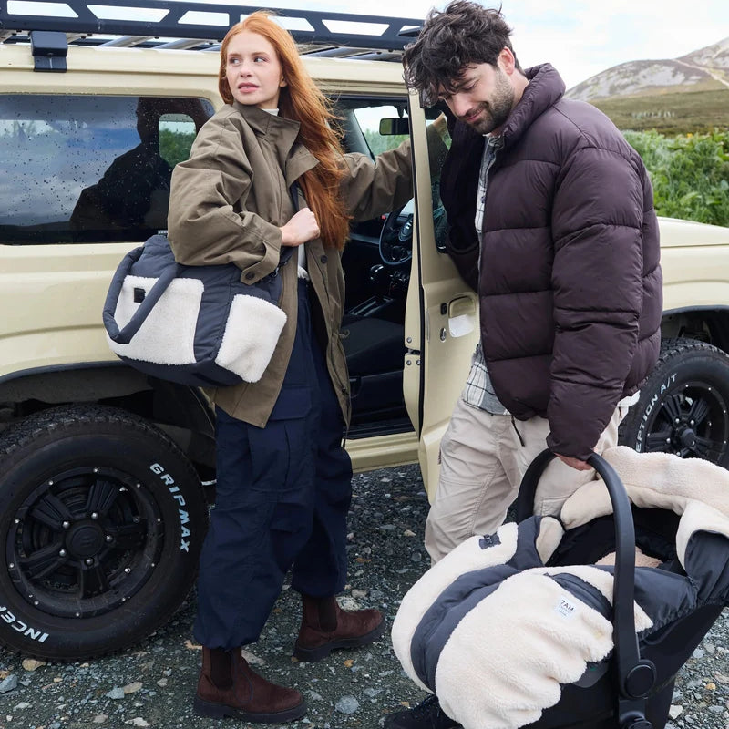 A man and a woman outdoors beside a beige off-road vehicle, each holding a matching black and cream fleece car seat liner set. The woman wears an olive green jacket and navy pants, and the man wears a dark brown puffer jacket and khaki pants.