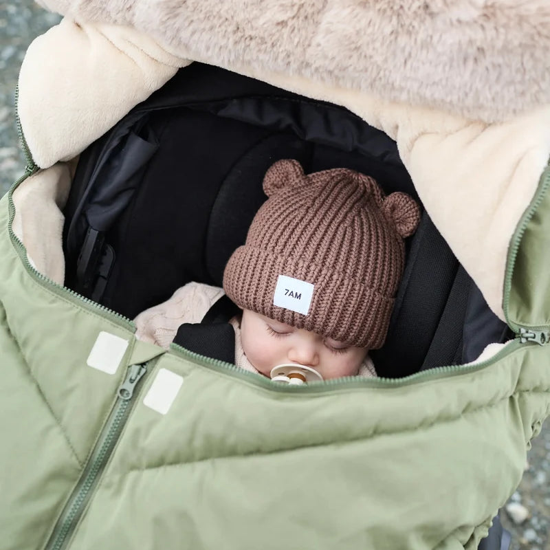 Baby sleeping peacefully in a moss brown Helsinki Car Seat Cocoon by 7AM Enfant with a plush faux-fur collar and matching knit bear-ear beanie.