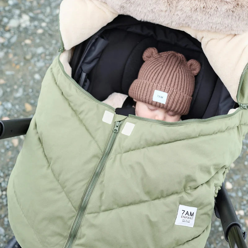 Baby sleeping peacefully in a moss brown Helsinki Car Seat Cocoon by 7AM Enfant with a plush faux-fur collar and matching knit bear-ear beanie.