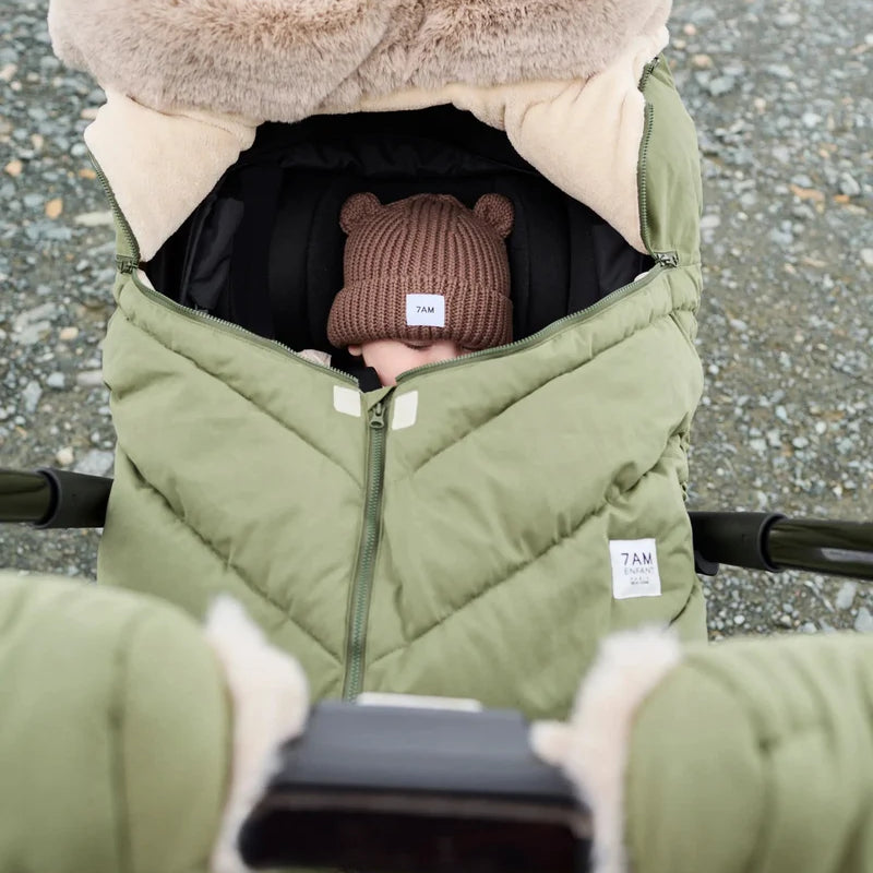 Baby sleeping peacefully in a moss brown Helsinki Car Seat Cocoon by 7AM Enfant with a plush faux-fur collar and matching knit bear-ear beanie.