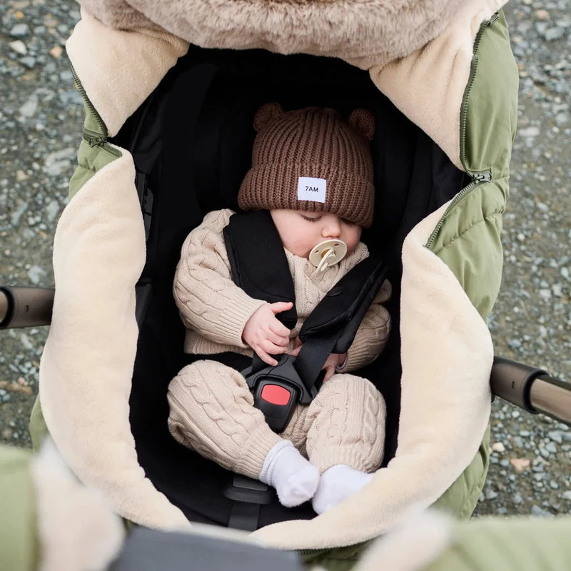 Baby sleeping peacefully in a moss brown Helsinki Car Seat Cocoon by 7AM Enfant with a plush faux-fur collar and matching knit bear-ear beanie.