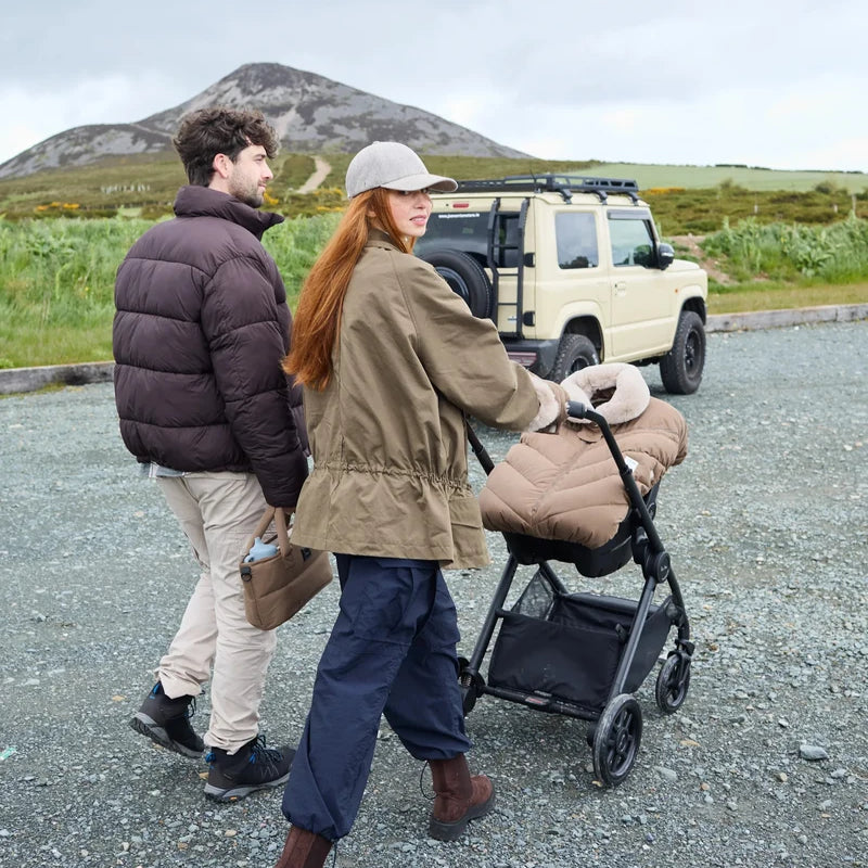 Lifestyle view of parents outdoors with a stroller featuring the moss brown Helsinki Car Seat Cocoon by 7AM Enfant parked near an off-road vehicle with a mountain landscape in the background.