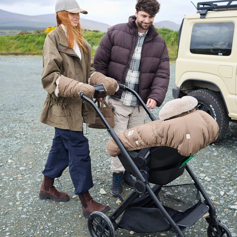 Lifestyle view of parents outdoors with a stroller featuring the moss brown Helsinki Car Seat Cocoon by 7AM Enfant parked near an off-road vehicle with a mountain landscape in the background.