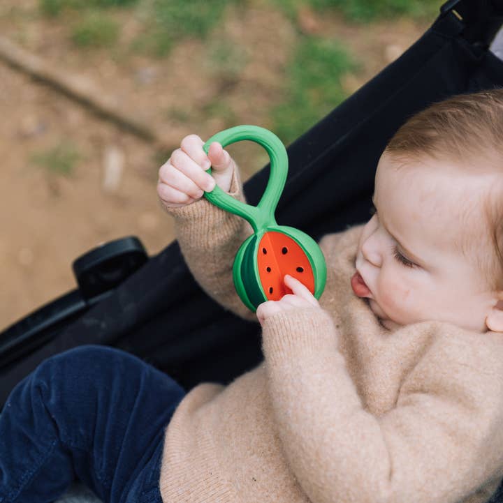 A baby in a beige sweater and blue pants holds a green and red watermelon-shaped teething toy while sitting in a black stroller outdoors.
