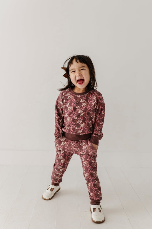 A young child wearing a matching long-sleeve sweatshirt and jogger pants set with a dusty rose and dark brown floral pattern, standing against a light gray studio background.