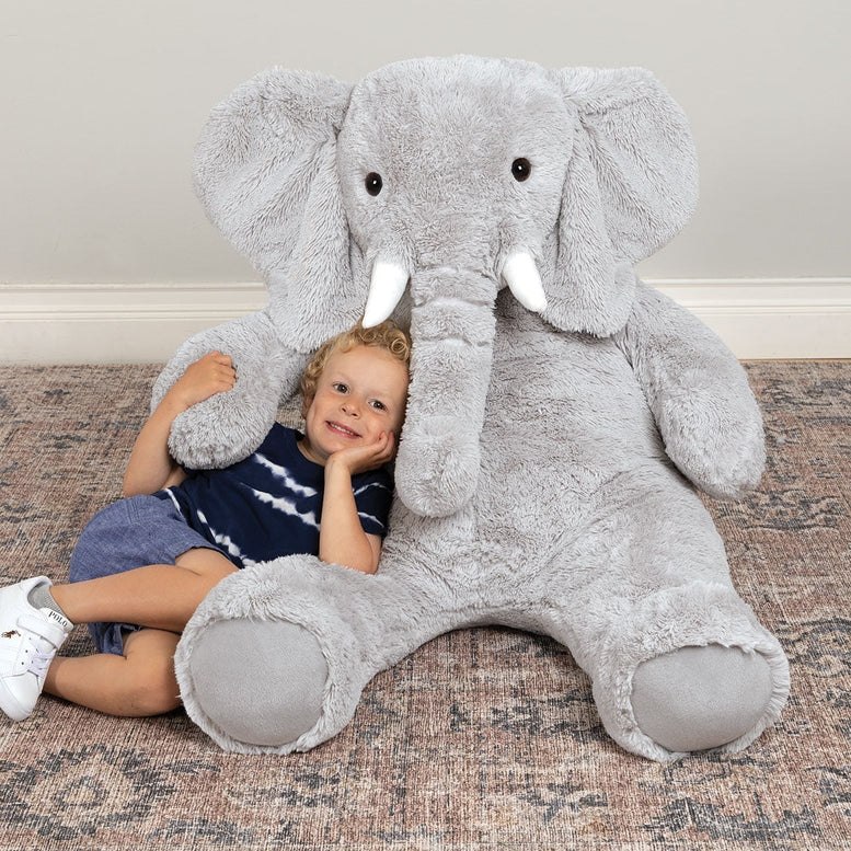 Child sitting on a large gray stuffed elephant toy on a carpeted floor.