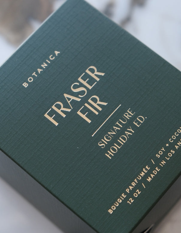 Green packaging of Fraser Fir candle with gold text on a blurred background