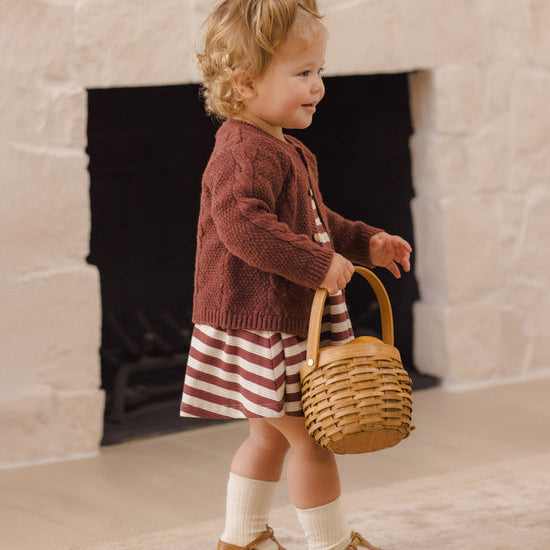 A toddler stands in front of a stone fireplace holding a small woven basket. The child wears a rust-brown knit cardigan over a red-and-white striped dress, white socks, and tan Mary Jane shoes.
