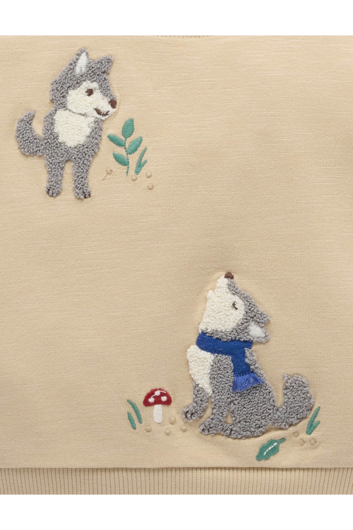 Close-up of a beige textile with two embroidered wolf motifs, one standing and one sitting with a blue scarf, plus small green leaves and a red-and-white mushroom detail.