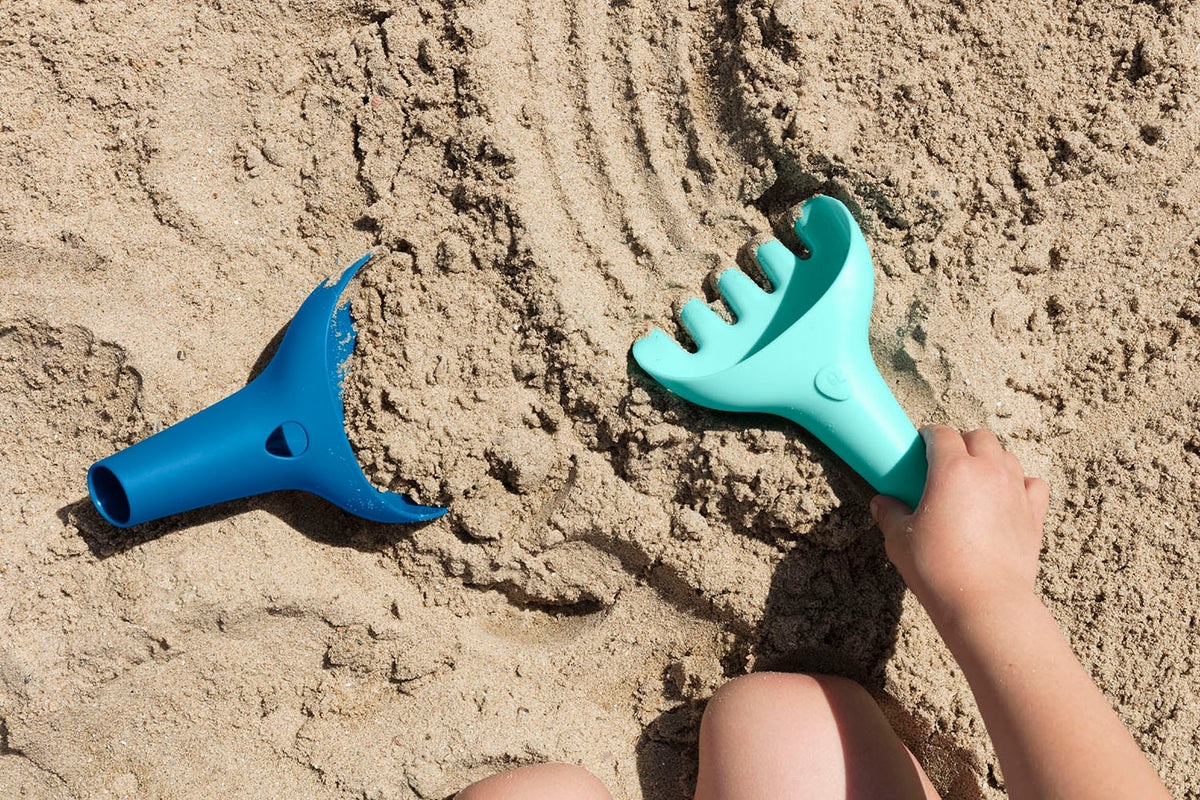Two blue sand toys on a sandy surface with a hand reaching towards one of them.
