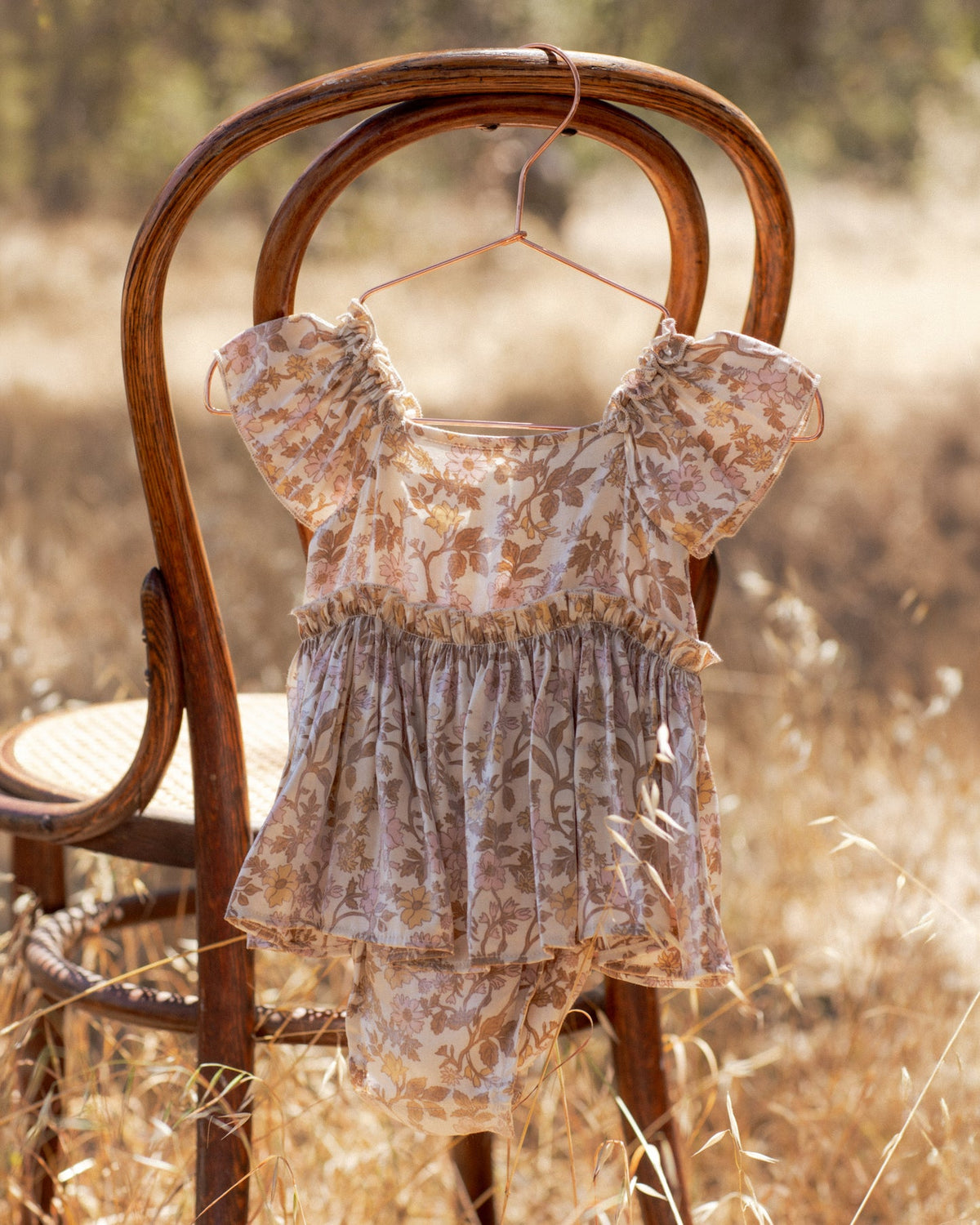 A baby girl's short-sleeve romper and matching shorts set in a beige and brown floral print, hanging on a wooden hanger from the back of a rustic wooden chair in a dry grass field.