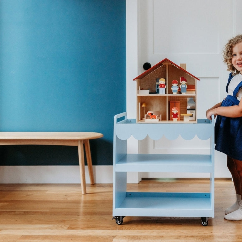 A young child with curly brown hair standing beside a blue rolling utility cart, reaching toward an open-faced wooden dollhouse by Mentari filled with miniature furniture.