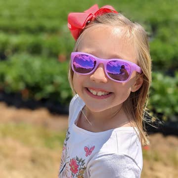 A young girl smiling in a strawberry field while wearing pink sunglasses with purple mirrored lenses and a red bow in her hair.
