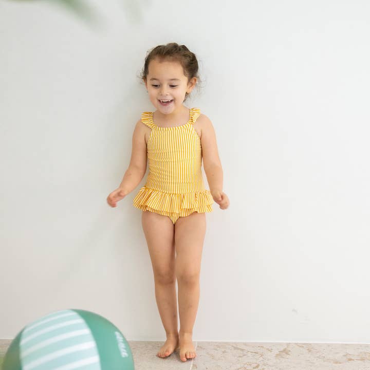 A young child smiling while wearing a yellow striped one-piece swimsuit, standing indoors next to a green and light blue striped beach ball.
