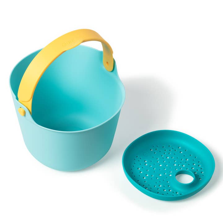 A light blue beach bucket with a yellow handle sits next to a matching darker teal round sand sifter with a handle hole on a white background.