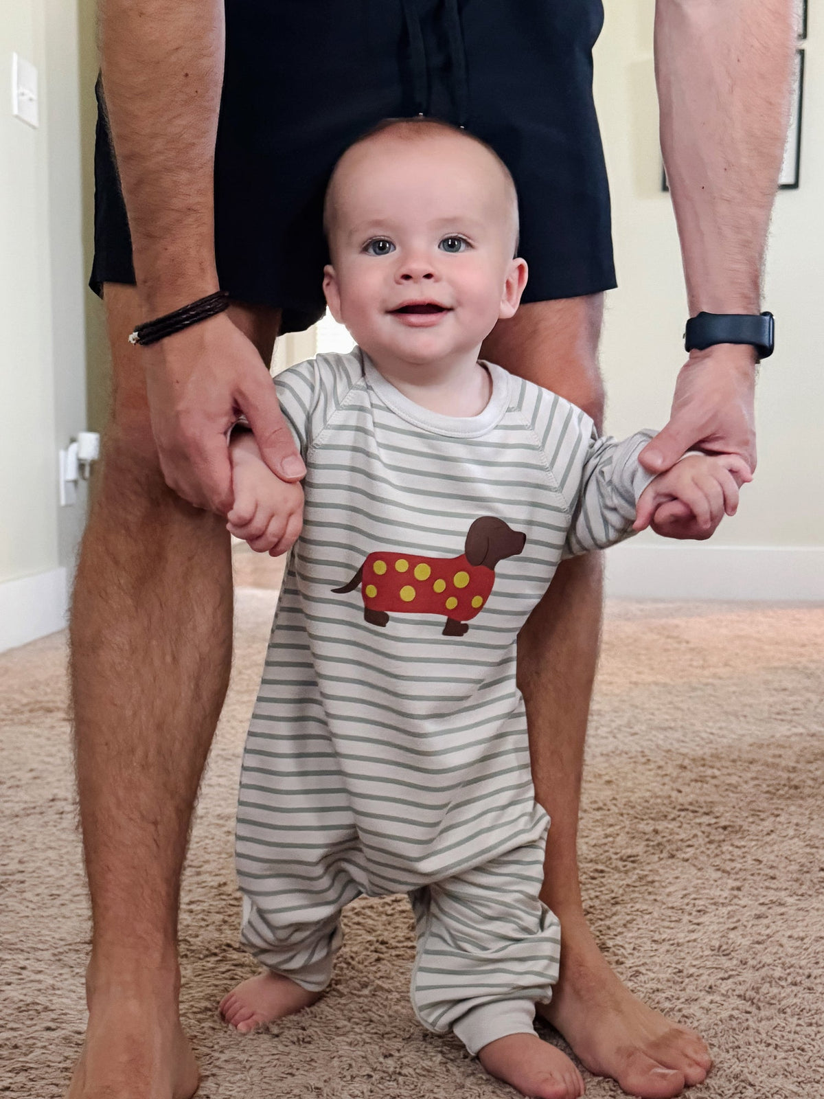 Baby in a striped onesie with a dachshund design, standing between two adults' legs.