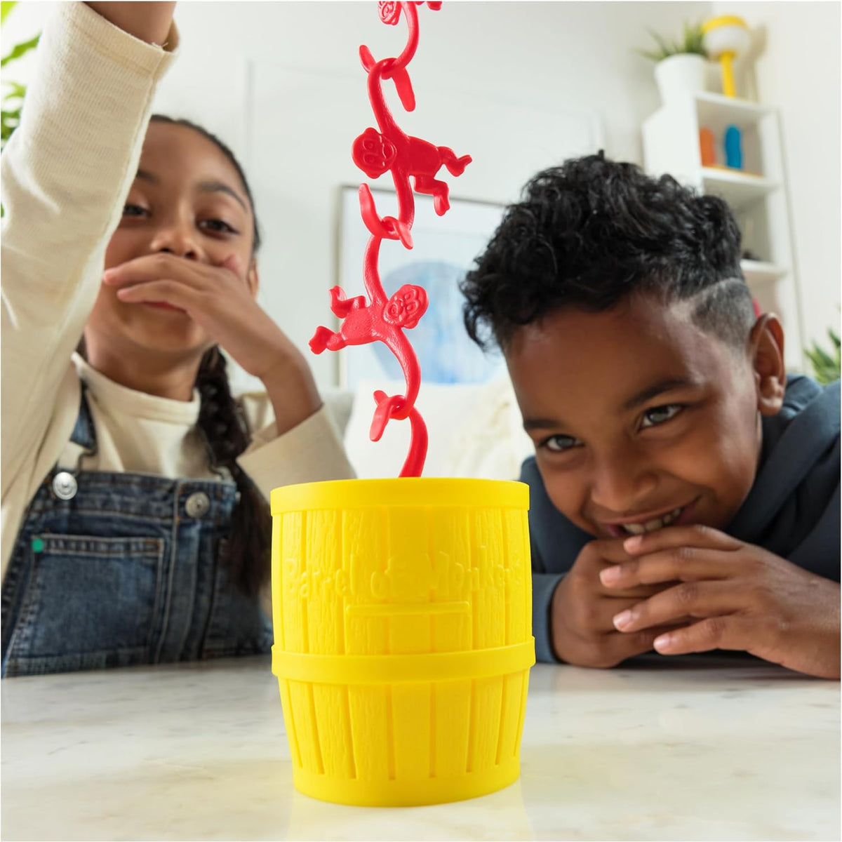 Two children playing with the Barrel of Monkeys Game by Continuum Games, linking a chain of red plastic monkeys above a yellow storage barrel.