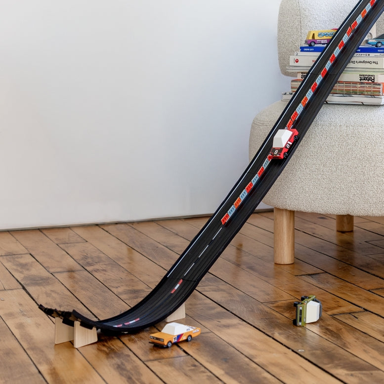Toy car track set on a wooden floor with books stacked on a chair in the background.