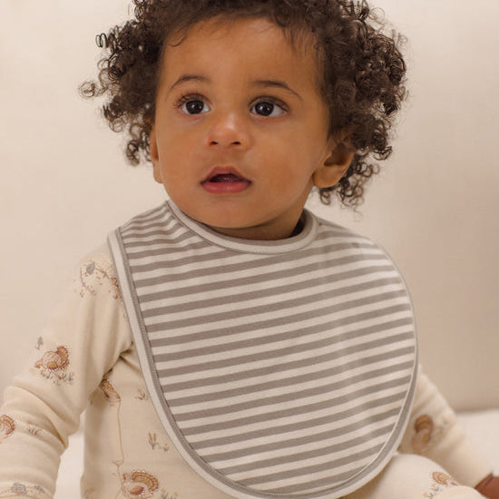 A close-up of a baby wearing a gray and white striped bib over a cream-colored patterned long-sleeve romper, set against a soft, warm off-white background.