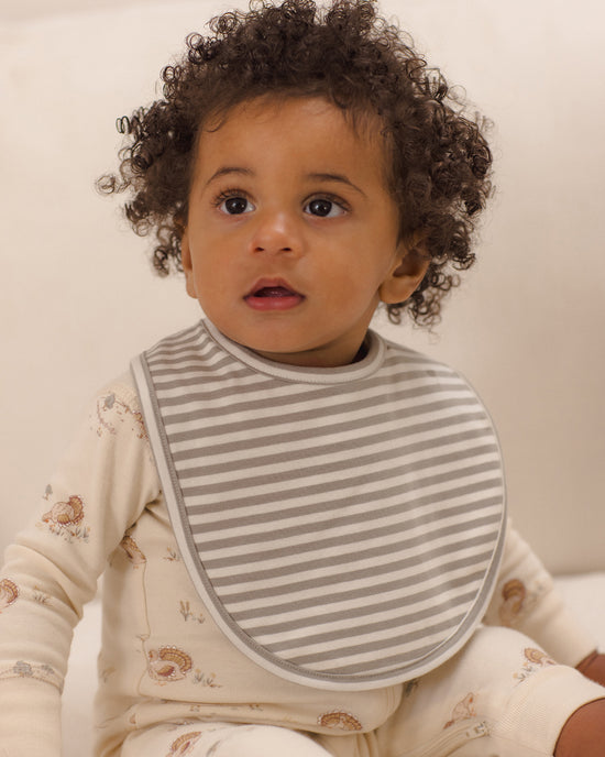 A close-up of a baby wearing a gray and white striped bib over a cream-colored patterned long-sleeve romper, set against a soft, warm off-white background.