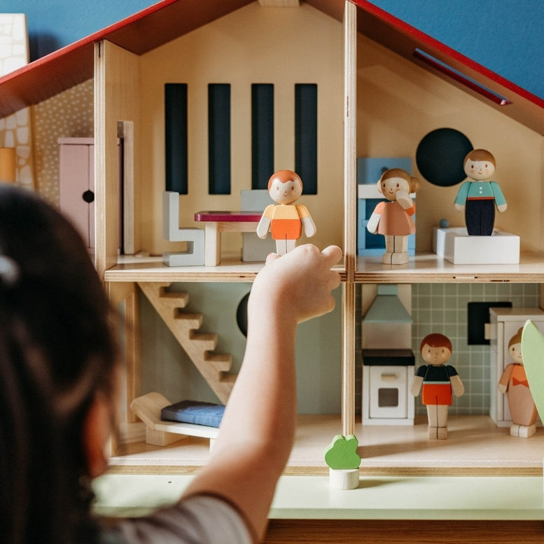 A child's hand reaches toward the open-front Wooden Cedar Chalet Modernist Dollhouse by Mentari, with miniature figures and furniture arranged across its six rooms and patio.